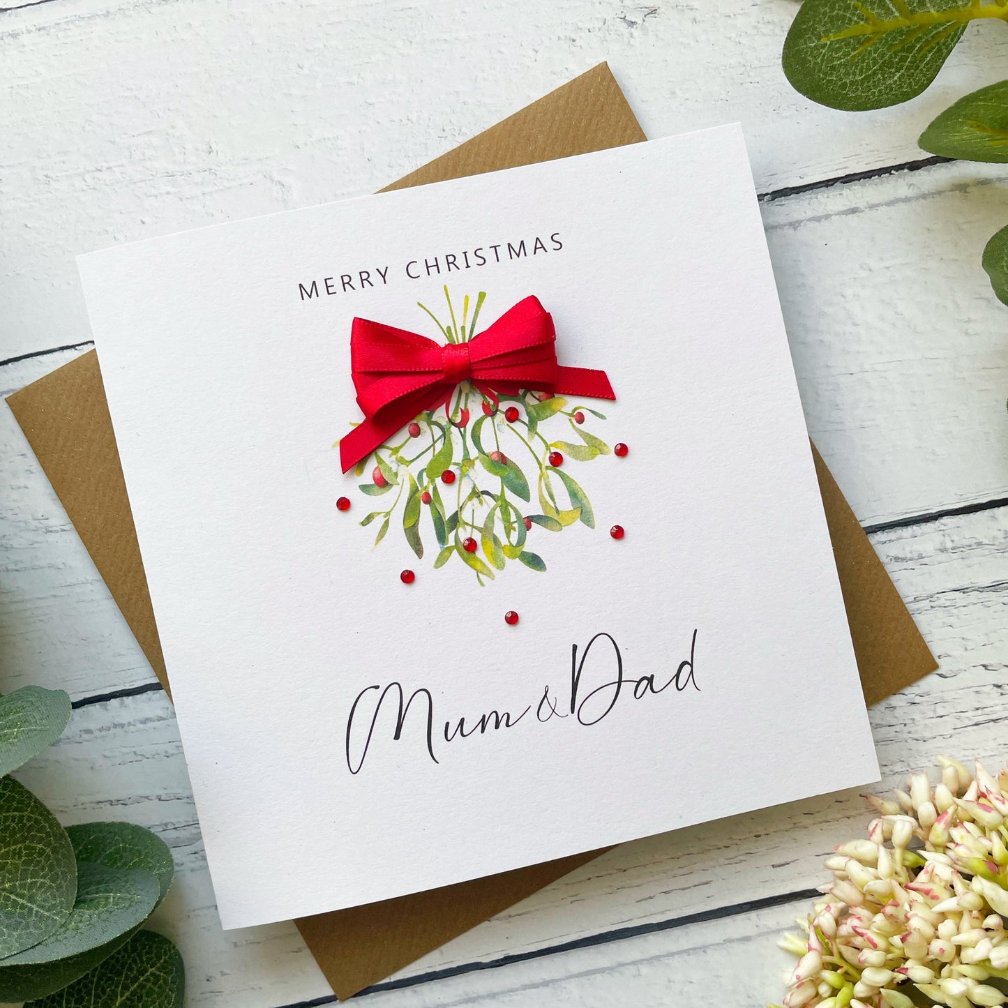 A square white Christmas greeting card addressed to Mum & Dad, featuring a watercolor mistletoe design with a red bow, placed on top of a brown envelope over a white wood-plank surface with green leaves and flowers.