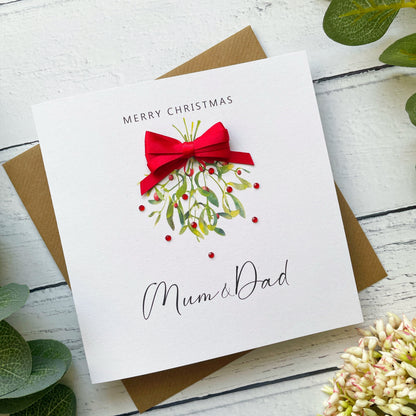 A square white Christmas greeting card addressed to Mum & Dad, featuring a watercolor mistletoe design with a red bow, placed on top of a brown envelope over a white wood-plank surface with green leaves and flowers.