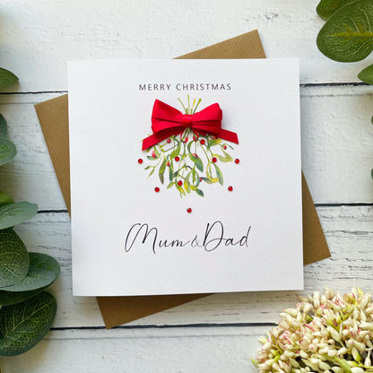 A square white Christmas greeting card with 'MERRY CHRISTMAS' at the top and 'Mum & Dad' in script at the bottom, featuring a red bow and mistletoe illustration. The card is placed on a brown envelope over a white wood surface with green leaves and pale flowers around it.