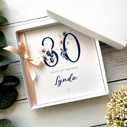 Gift box with a birthday card inside, featuring the number 80 and the name of the recipient  on a white wooden surface.
