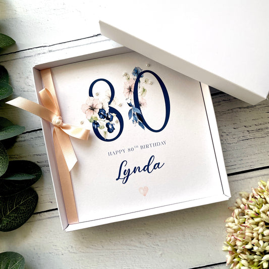 Gift box with a birthday card inside, featuring the number 80 and the name of the recipient  on a white wooden surface.