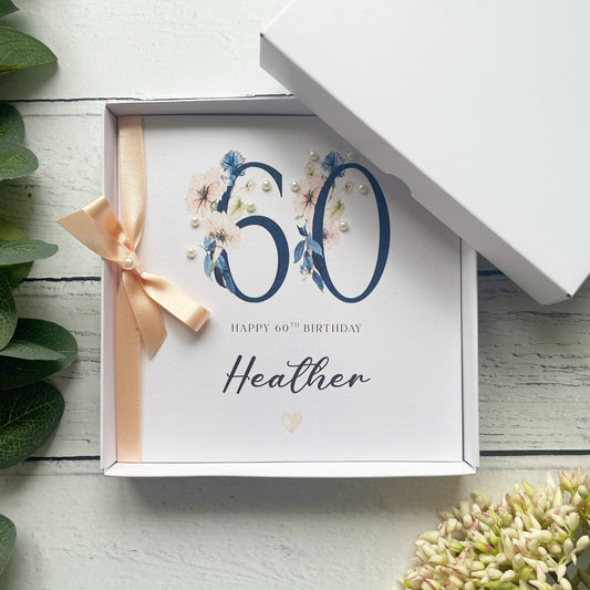 60th birthday card with floral design and ribbon in a box on a white wooden surface.