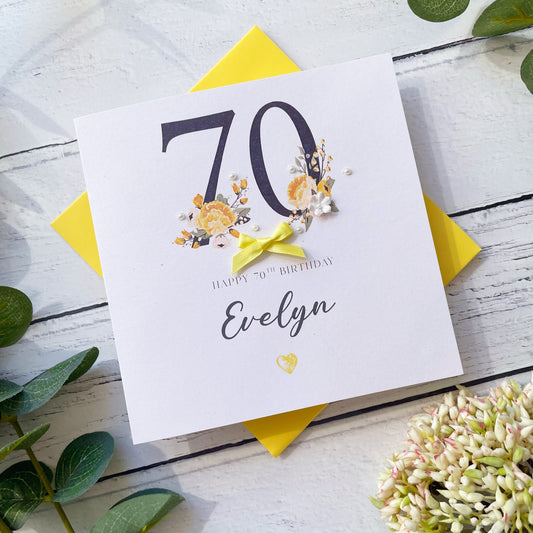 70th birthday card for personalised with name and age, with bright yellow bow on a white wooden surface with yellow envelope and floral decorations.
