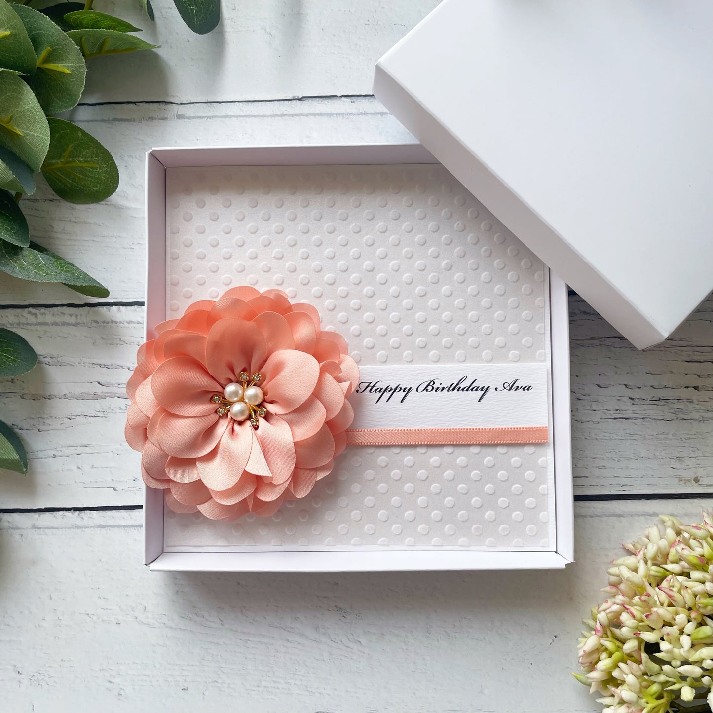 Birthday Card in a Gift box with a peach flower embellishment and personalised text details, on a white wooden surface.