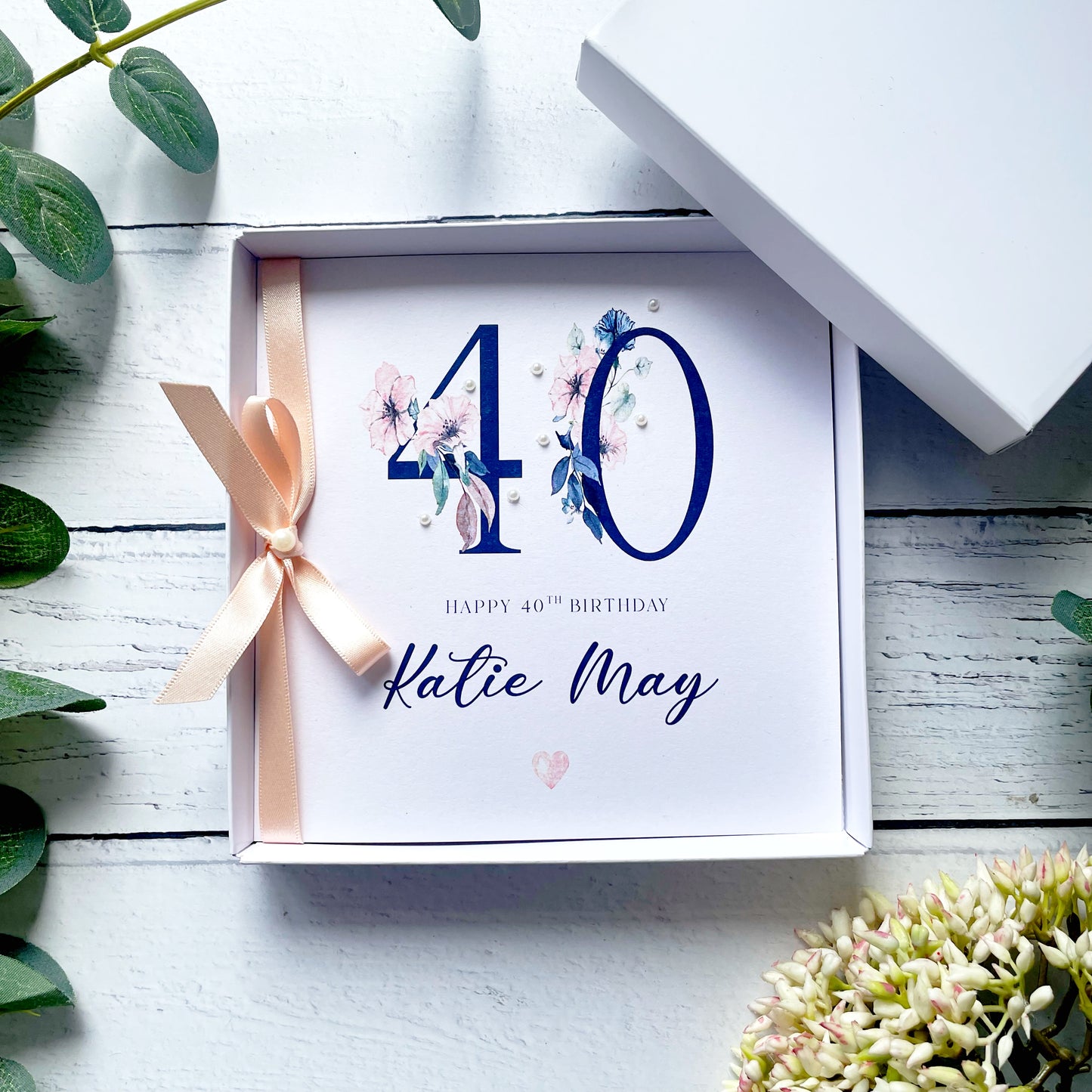 Boxed birthday card with '40' and 'Happy 40th Birthday Katie May' text on a white wooden surface with greenery.