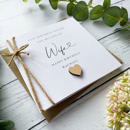 Greeting card with 'Last birthday before you become my wife' (name) text, tied with twine on a white wooden surface with greenery.