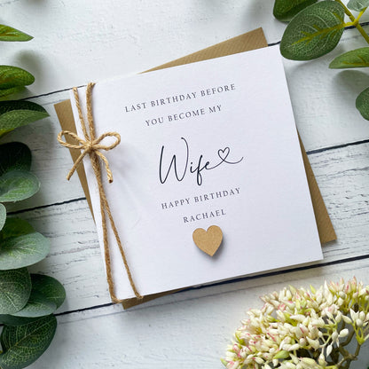 Greeting card with 'Last birthday before you become my wife' (name) text, tied with twine on a white wooden surface with greenery.