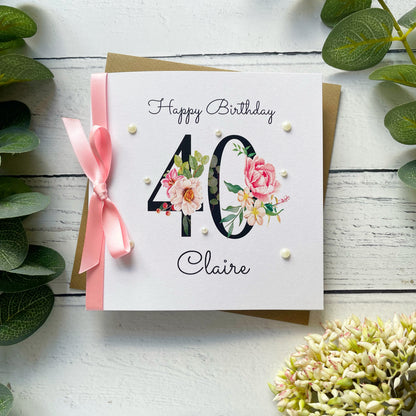 Birthday card with floral design and number 40, featuring a pink ribbon on a white wooden background.