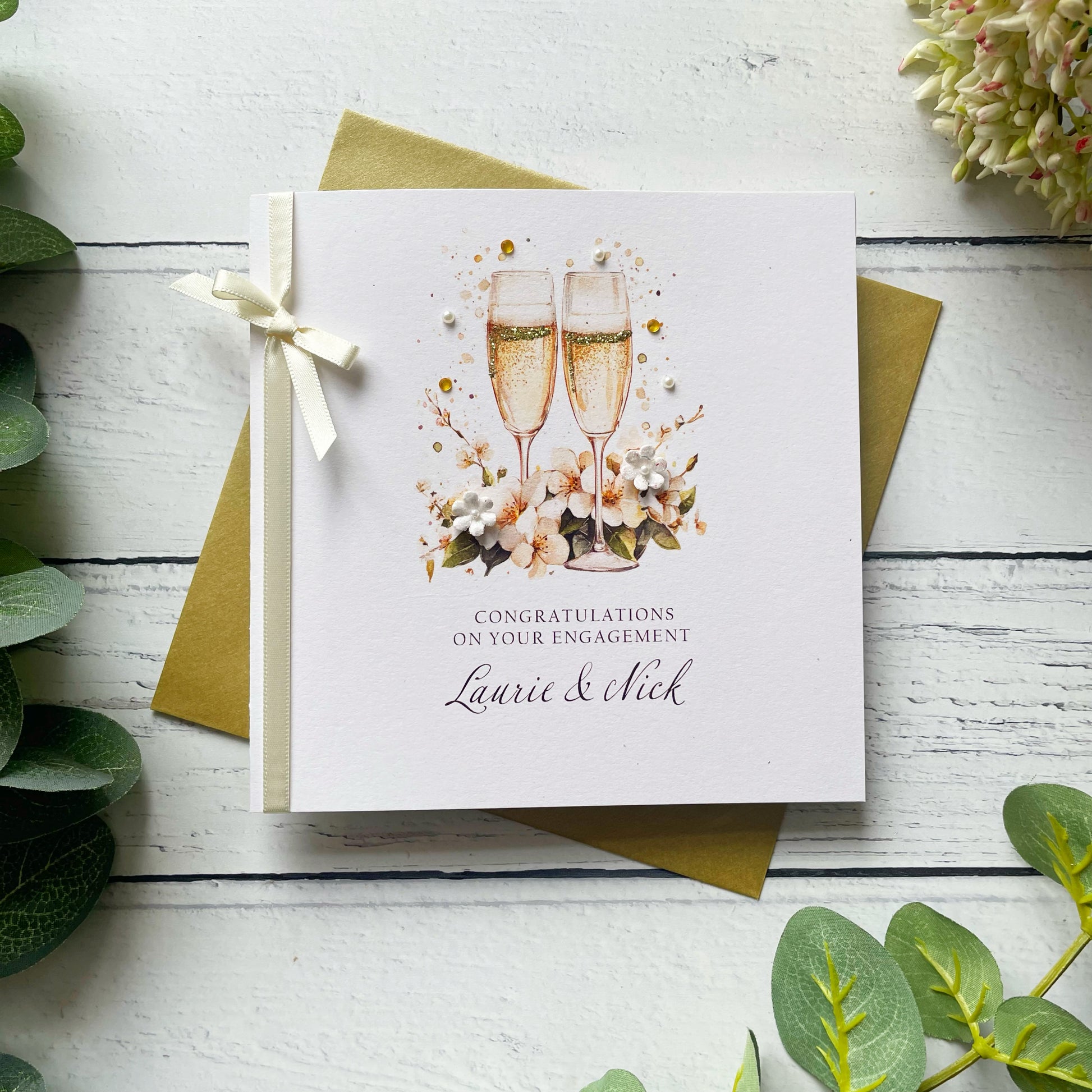Engagement card with champagne glasses and floral design on a white wooden surface with greenery.