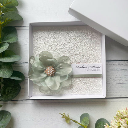 Decorative box with a floral design wedding card with personalised text. With a pearl brooch on a white wooden surface.