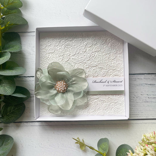 Decorative box with a floral design wedding card with personalised text. With a pearl brooch on a white wooden surface.