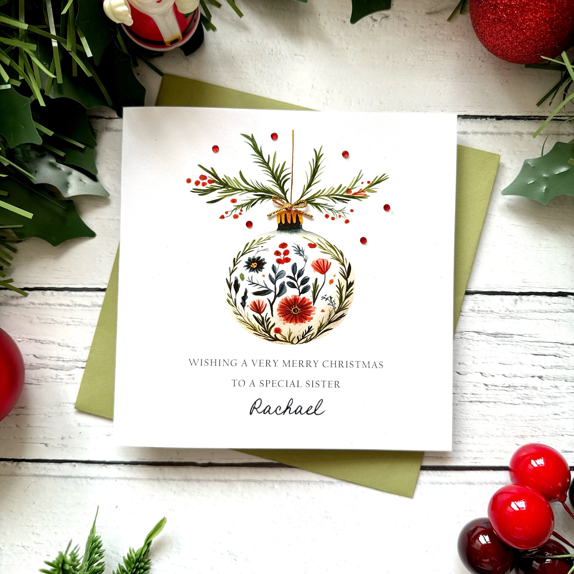 A square white Christmas card featuring a watercolour floral bauble with red poppies, dark botanicals and green foliage, topped with pine sprigs, red berries and a tiny gold bow, dotted with red gem embellishments. Text reads "Wishing a Very Merry Christmas to a Special Sister" in spaced capitals with "Rachael" in cursive script below. Displayed on a green envelope with holly and red berry sprigs.