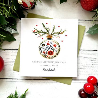 A square white Christmas card featuring a watercolour floral bauble with red poppies, dark botanicals and green foliage, topped with pine sprigs, red berries and a tiny gold bow, dotted with red gem embellishments. Text reads "Wishing a Very Merry Christmas to a Special Sister" in spaced capitals with "Rachael" in cursive script below. Displayed on a green envelope with holly and red berry sprigs.
