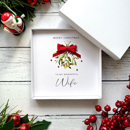 A white square gift box with the lid partly open, displaying a Christmas greeting card that reads 'MERRY CHRISTMAS TO MY WONDERFUL Wife' with a red bow and mistletoe illustration. The box is styled on a white wood surface with holiday greenery, red berries, and a small Santa figurine.