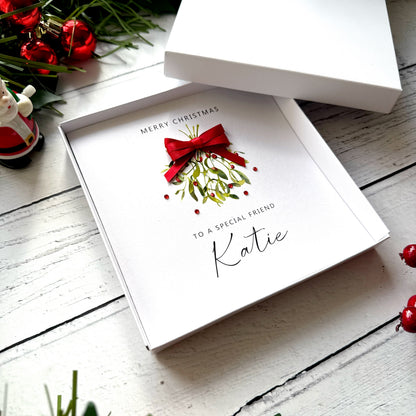 A white Christmas greeting card in a white gift box, featuring a watercolor illustration of mistletoe with a red bow and the text 'Merry Christmas to a special friend Katie'. The box is open on a white wooden surface with holiday decorations in the background.