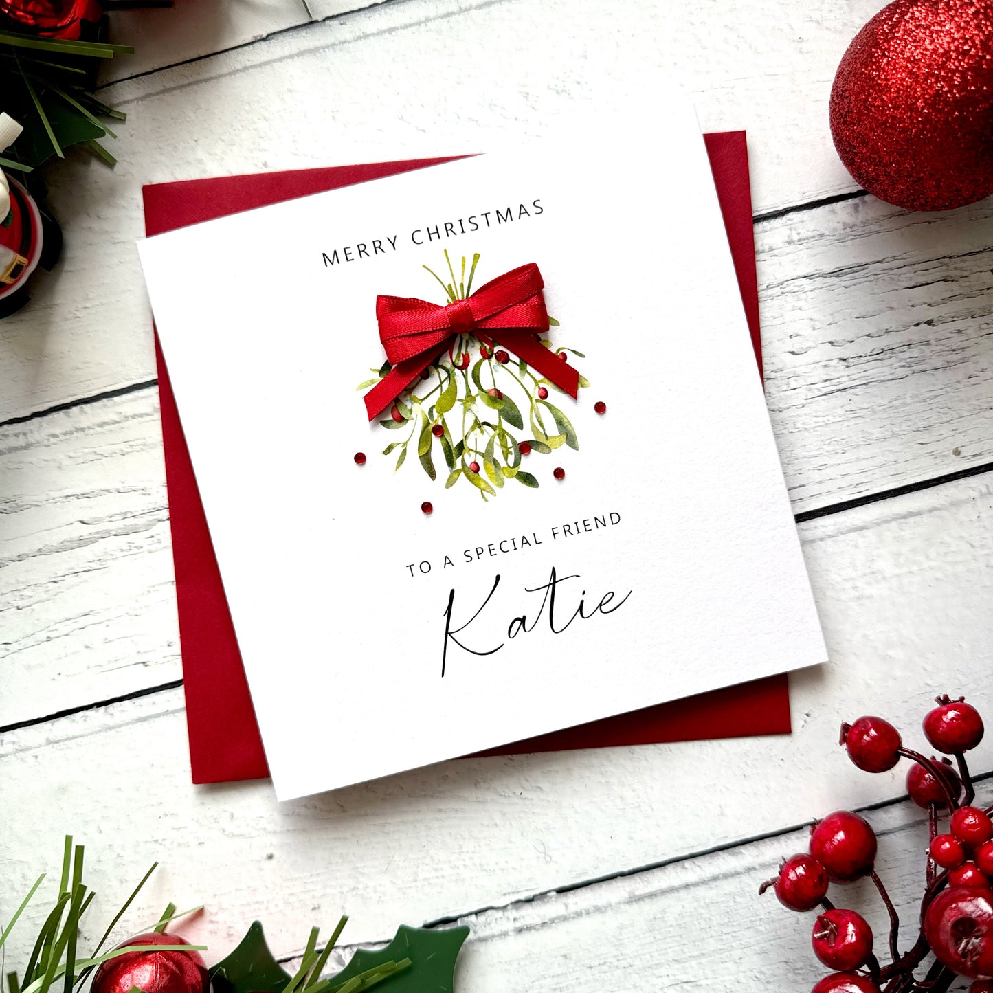 A square white Christmas greeting card with a red envelope, styled on a white wood surface with holiday decorations. The card reads 'MERRY CHRISTMAS TO A SPECIAL FRIEND Katie' and features an illustration of mistletoe with a red bow.