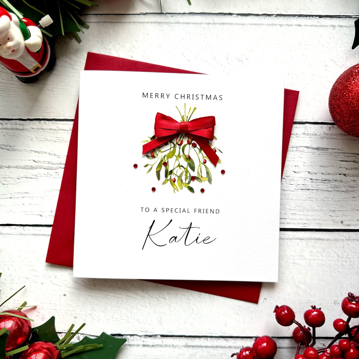 A square white Christmas card featuring a watercolour illustration of a mistletoe bunch with green leaves and red berries, tied with a real red satin ribbon bow and accented with small red gem embellishments scattered around the design. The text "Merry Christmas" is printed in small spaced capitals at the top of the card, followed by "to a Special Friend" in small capitals and the name "Katie" in large elegant black cursive script below the illustration. The card sits on a matching red envelope on a white p