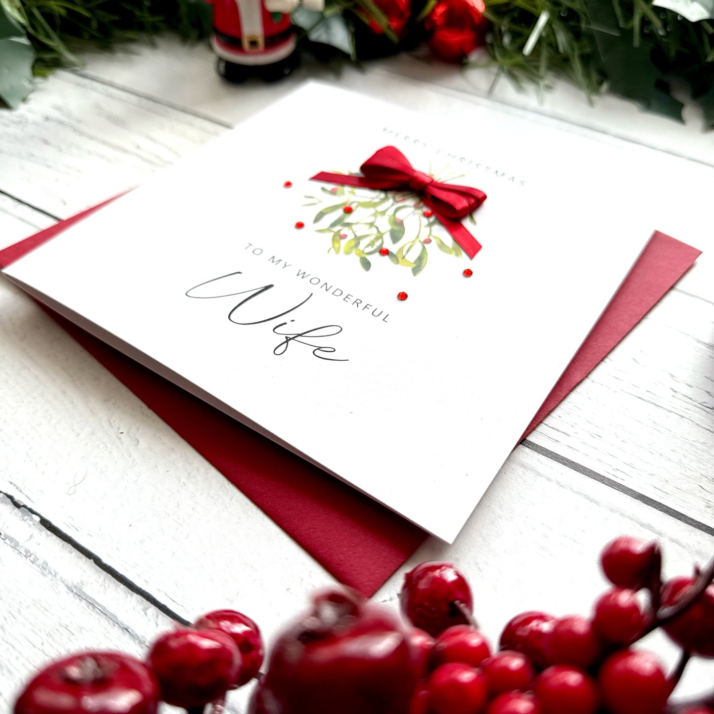 A white Christmas greeting card addressed to a wife, decorated with a red bow and mistletoe illustration, resting on a dark red envelope over a white wood surface with holiday decorations around it.