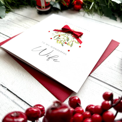 A white Christmas greeting card addressed to a wife, decorated with a red bow and mistletoe illustration, resting on a dark red envelope over a white wood surface with holiday decorations around it.