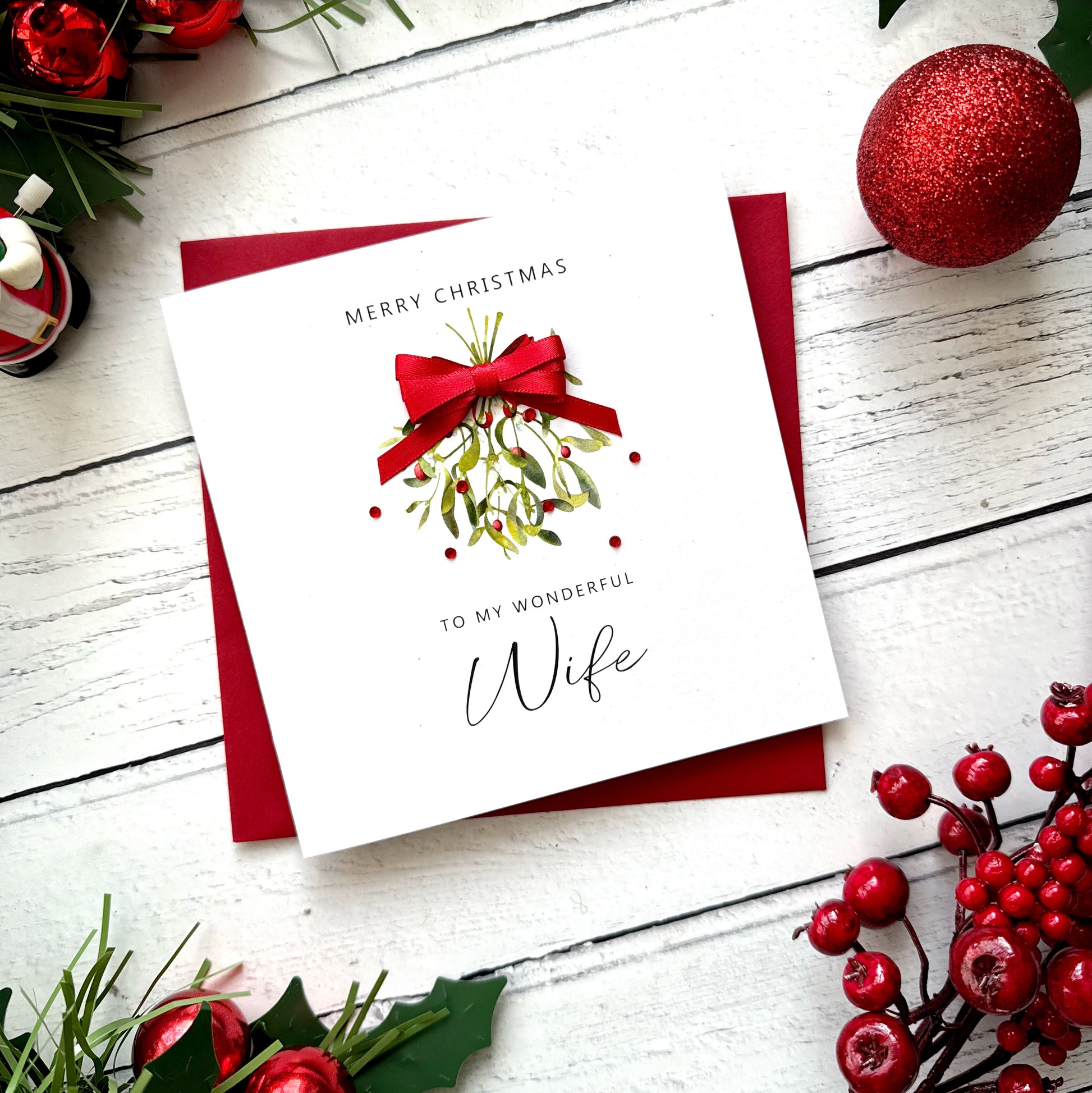 A square white Christmas greeting card with a red envelope, styled on a white wood surface with holiday decorations including red berries, green leaves, and a red ornament.