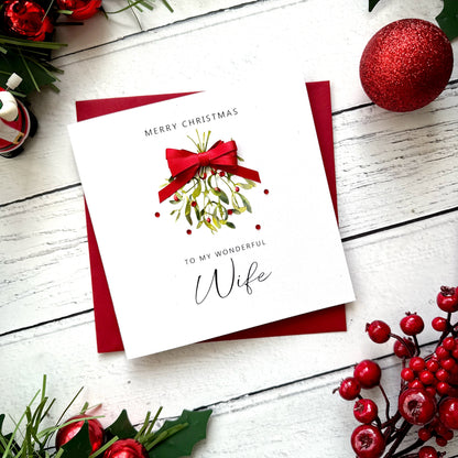 A square white Christmas greeting card with a red envelope, styled on a white wood surface with holiday decorations including red berries, green leaves, and a red ornament.
