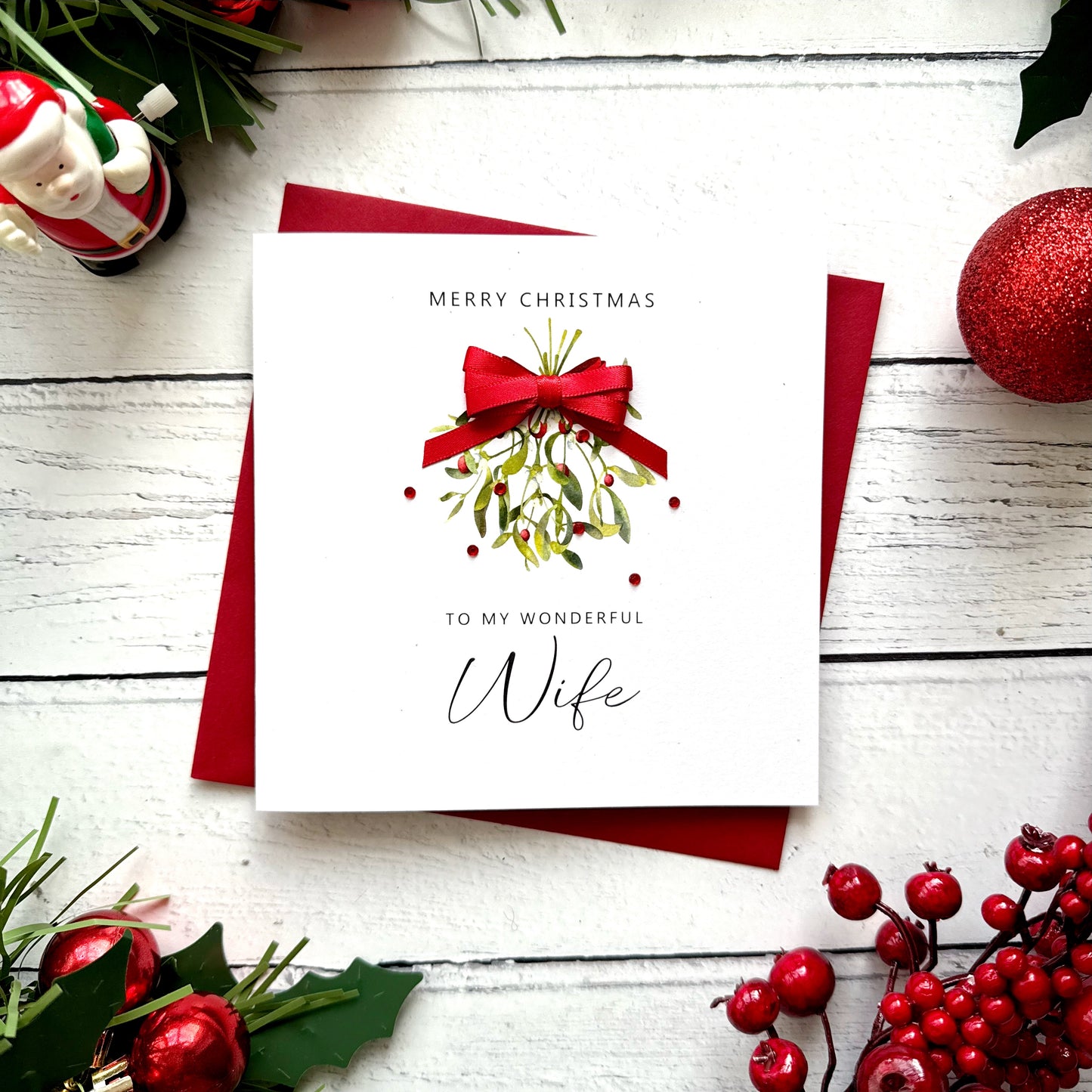 A square white Christmas card featuring a watercolour illustration of a mistletoe bunch with green leaves and red berries, tied with a real red satin ribbon bow and accented with small red gem embellishments scattered around the design. The text "Merry Christmas" is printed in small spaced capitals at the top of the card, with "to my Wonderful" in small capitals and "Wife" in large elegant black cursive script below the illustration. The card sits on a matching red envelope on a white painted wooden surface