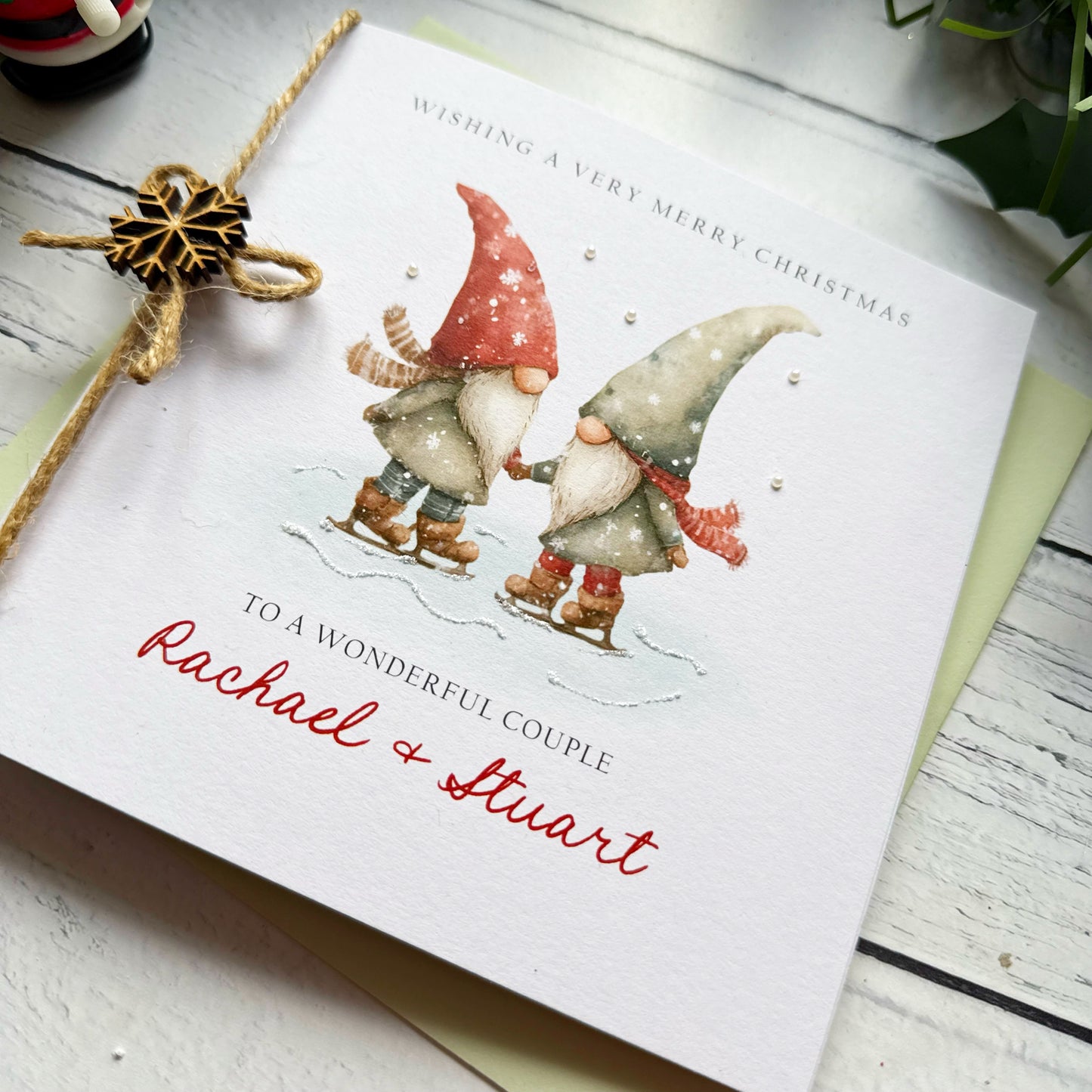 Christmas card with two gnomes ice skating, personalised for a special couple with names. Decorated with glitter, pearls, twine and a snowflake on a white surface.