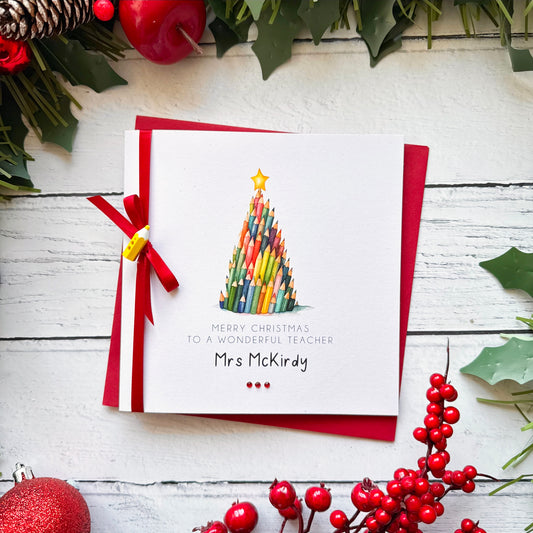 A personalised Christmas greeting card featuring a colourful pencil Christmas tree and the text 'Merry Christmas to a wonderful teacher Mrs McKirdy', styled on a white wooden background with red envelopes, holly, pinecones, and red berries.