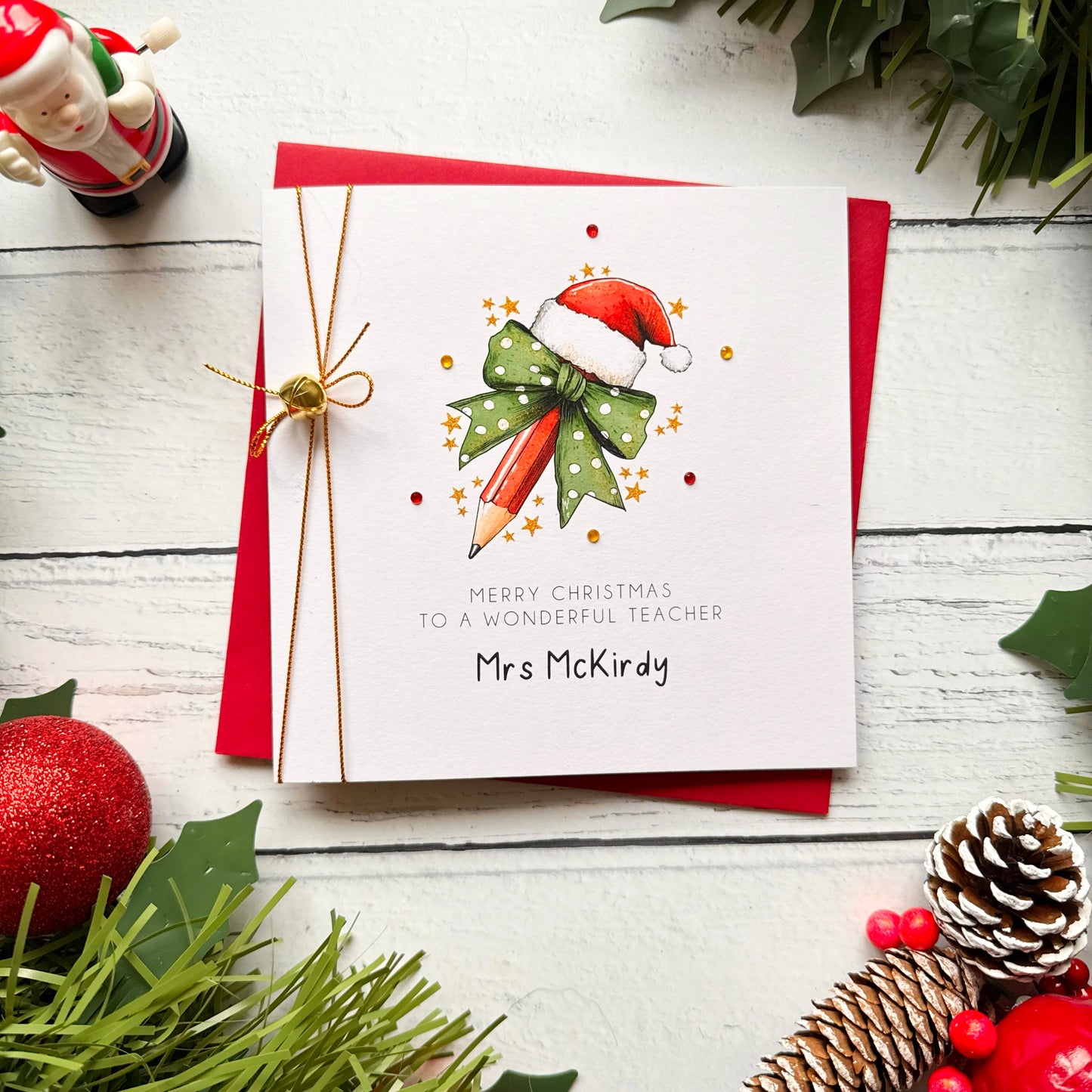 A personalised Christmas greeting card for a teacher, featuring a watercolor illustration of a pencil in a Santa hat with a green polka-dot bow. The card is personalized with the name 'Mrs McKirky' and tied with a gold string, displayed on a red envelope over a white wooden background decorated with holiday greenery and ornaments.