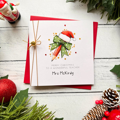 A personalised Christmas greeting card for a teacher, featuring a watercolor illustration of a pencil in a Santa hat with a green polka-dot bow. The card is personalized with the name 'Mrs McKirky' and tied with a gold string, displayed on a red envelope over a white wooden background decorated with holiday greenery and ornaments.