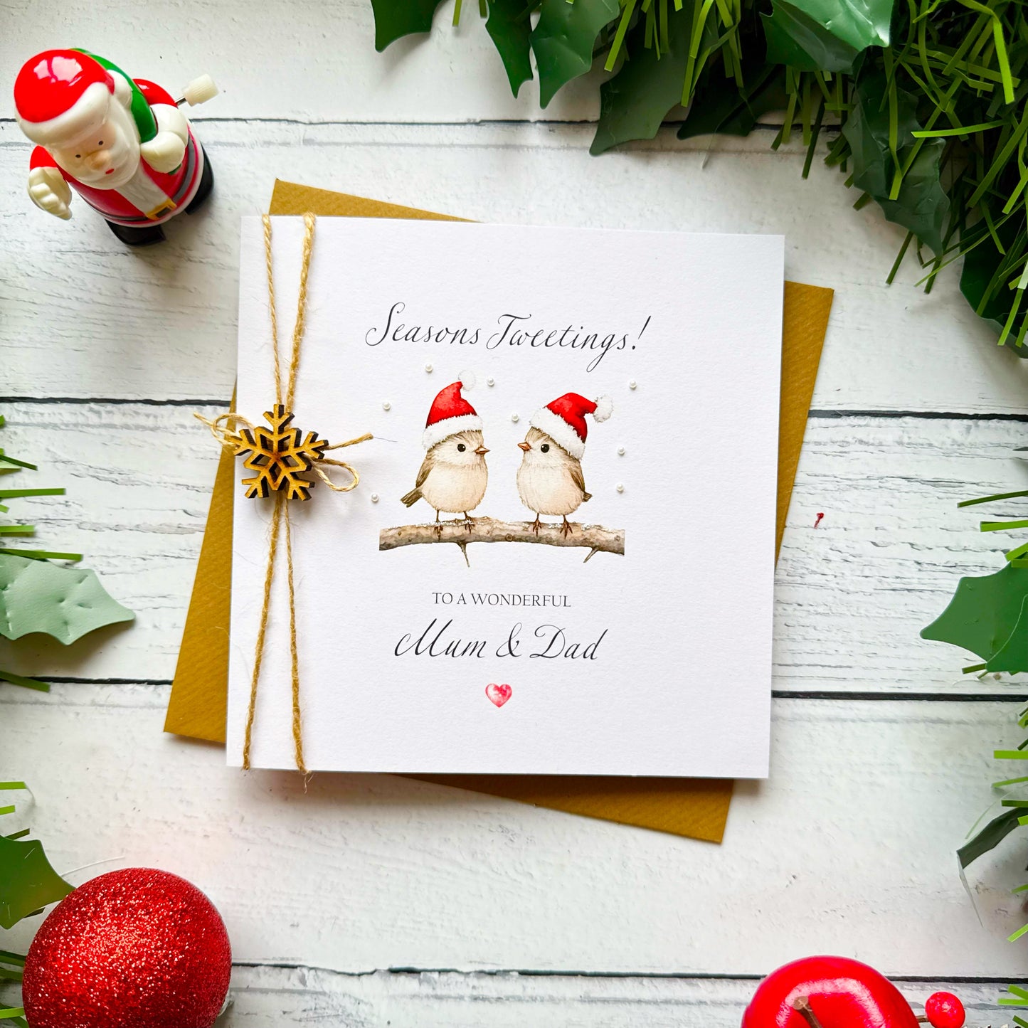 A square white Christmas card featuring two fluffy birds in red Santa hats perched on a snow-dusted branch with tiny pearl embellishments scattered around. Text reads "Seasons Tweetings!" in cursive script, "to a Wonderful" in small capitals and "Mum & Dad" in elegant cursive, finished with a small red heart. Tied with natural twine and a wooden snowflake charm on a brown kraft envelope.