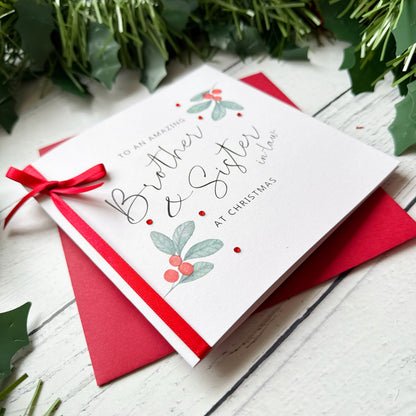 A white greeting card with elegant black script reading 'To an amazing Brother & Sister at Christmas' and small holly illustrations, tied with a red ribbon and placed on a red envelope over a white wood surface with holly leaves around it.