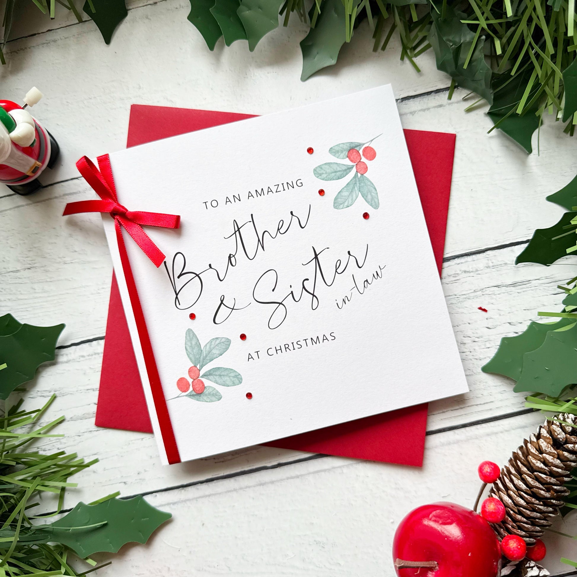 A white Christmas greeting card addressed 'To an Amazing Brother & Sister in Law at Christmas' with red berries and holly illustrations, tied with a red ribbon and placed on a red envelope over a white wood surface with holiday greenery and props.