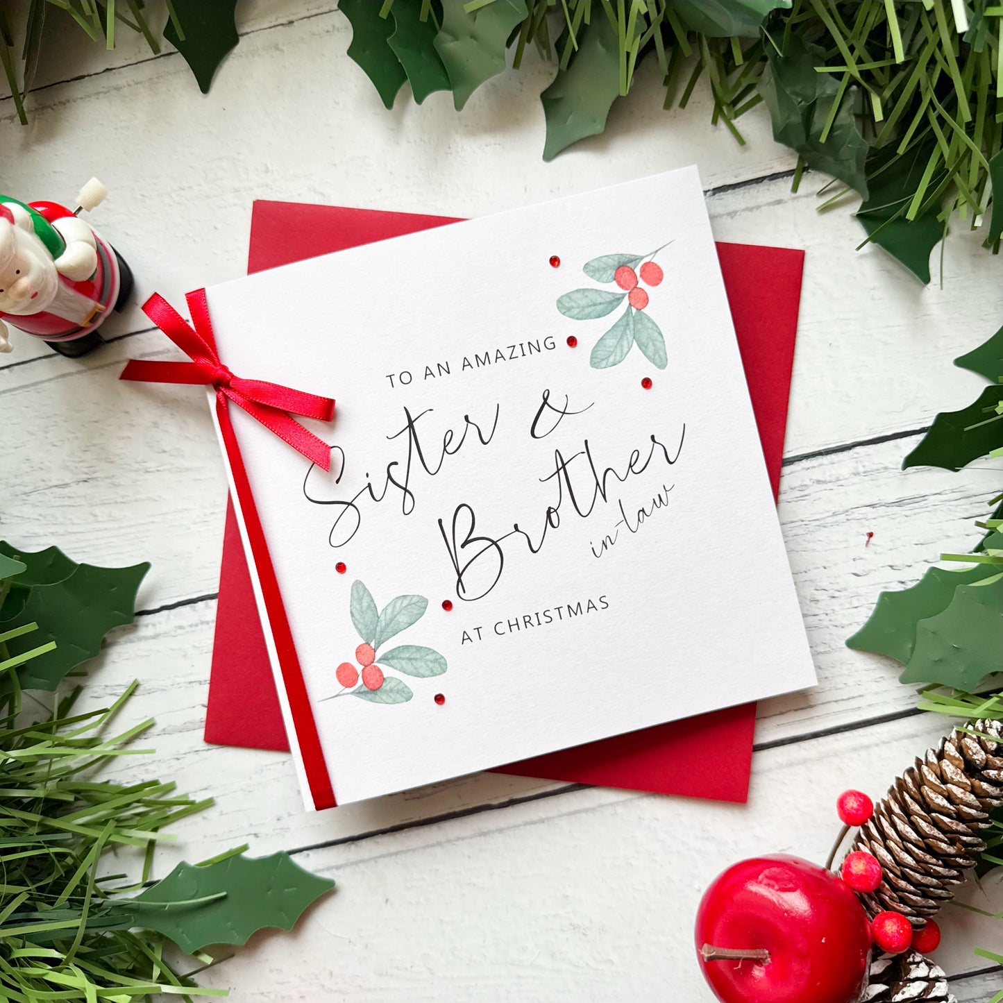 A square Christmas greeting card with a red ribbon and red envelope, styled on a white wooden surface with holly leaves, pine branches, a small figurine, and red berries. The card reads, 'TO AN AMAZING Sister & Brother in law AT CHRISTMAS'.