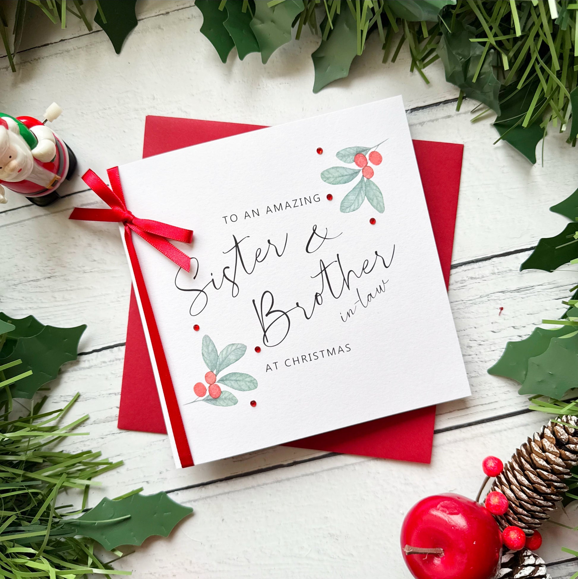A square Christmas greeting card with a red ribbon and red envelope, styled on a white wooden surface with holly leaves, pine branches, a small figurine, and red berries. The card reads, 'TO AN AMAZING Sister & Brother in law AT CHRISTMAS'.