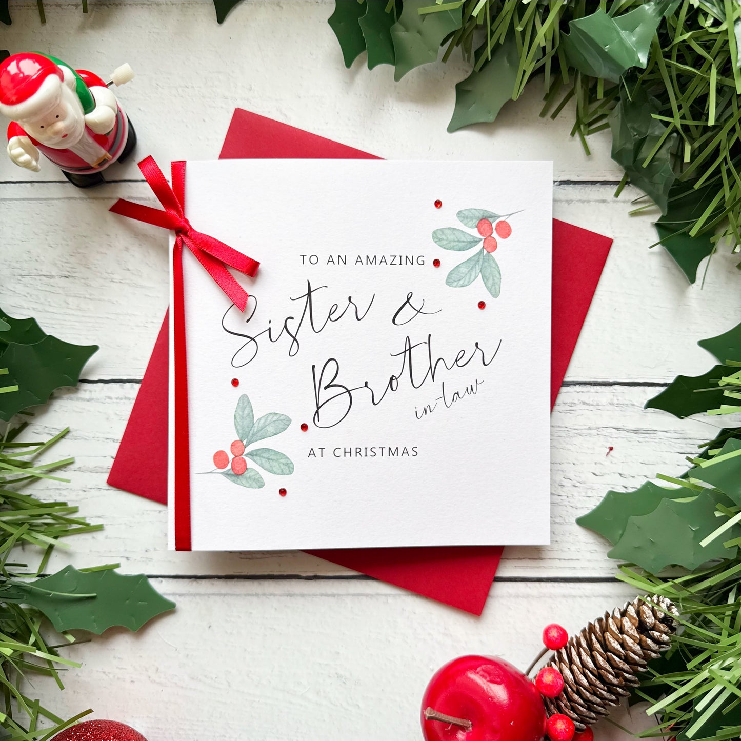 A square white Christmas greeting card with elegant black script reading 'To an amazing Sister & Brother in law! AT CHRISTMAS' and small holly and berry illustrations, tied with a red ribbon and placed on red envelopes. The card is styled in a flat lay with holly leaves, pinecones, red apples, a Santa figurine, and a white wood plank background.
