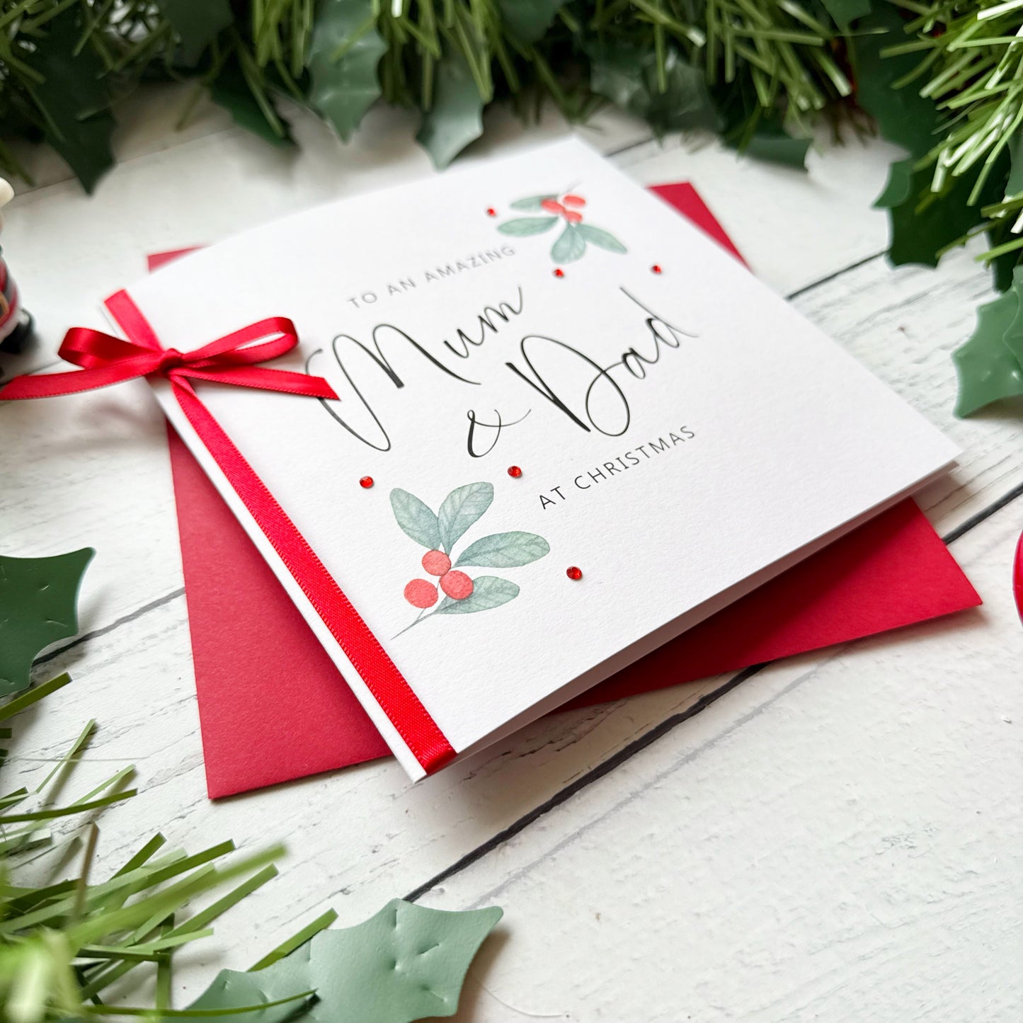 A white Christmas greeting card addressed to Mum and Dad, decorated with a red ribbon and bow, red berries, and green holly leaves. The card is placed on top of a red envelope on a white wooden surface, surrounded by green pine garlands.