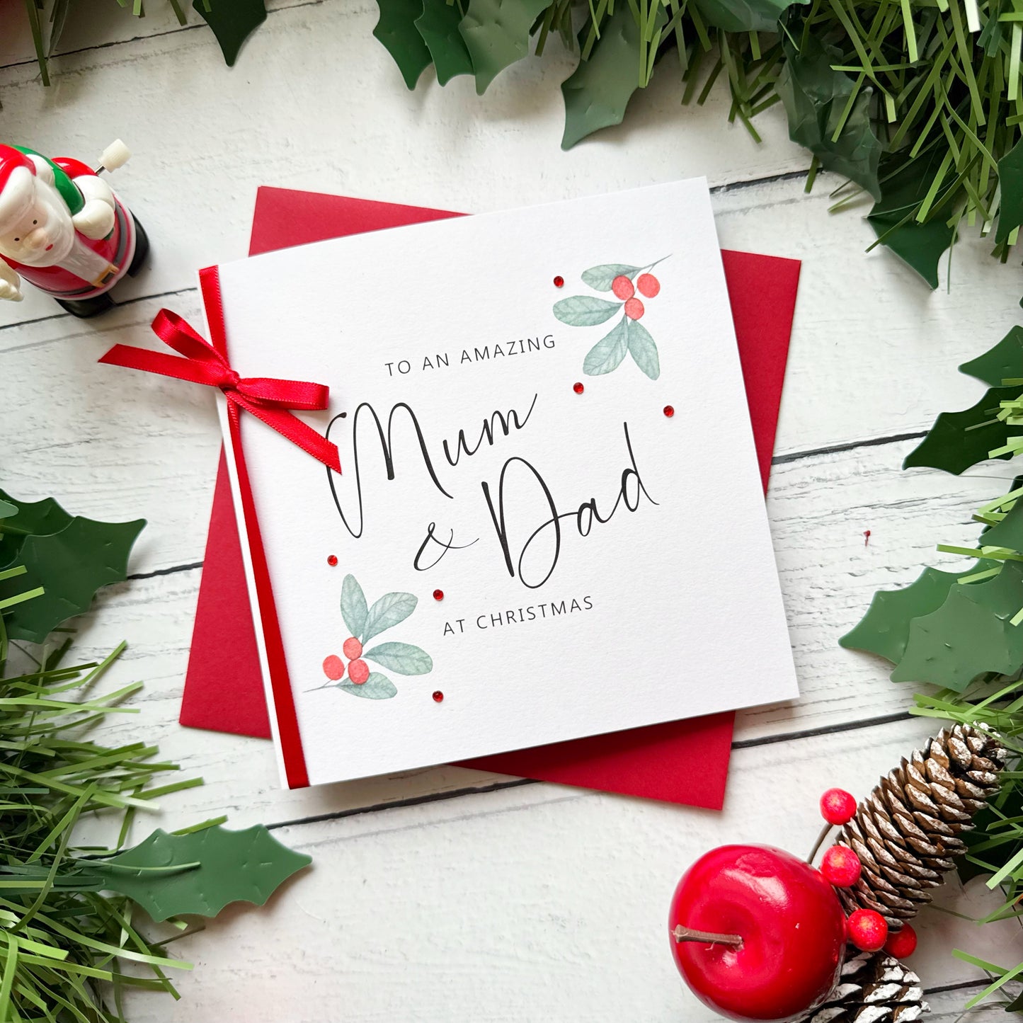 A square white Christmas card featuring watercolour holly berry sprigs with sage green leaves and red berries in the upper right and lower left corners, accented with small scattered red gem embellishments. The text reads "to an Amazing" in small spaced capitals at the top, with "Mum & Dad" in large flowing black cursive script in the centre, and "at Christmas" in small spaced capitals below. The card is tied with a real red satin ribbon bow on the left side and sits on a matching red envelope on a white pa