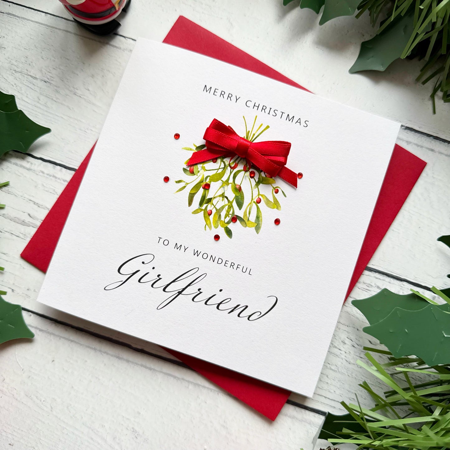 A square white Christmas card featuring a watercolour illustration of a mistletoe bunch with green leaves and red berries, tied with a real red satin ribbon bow and accented with small red gem embellishments scattered around the design. The text "Merry Christmas" is printed in small spaced capitals at the top of the card, with "to my Wonderful" in small capitals and "Girlfriend" in large elegant black cursive script below the illustration. The card sits on a matching red envelope on a white painted wooden s