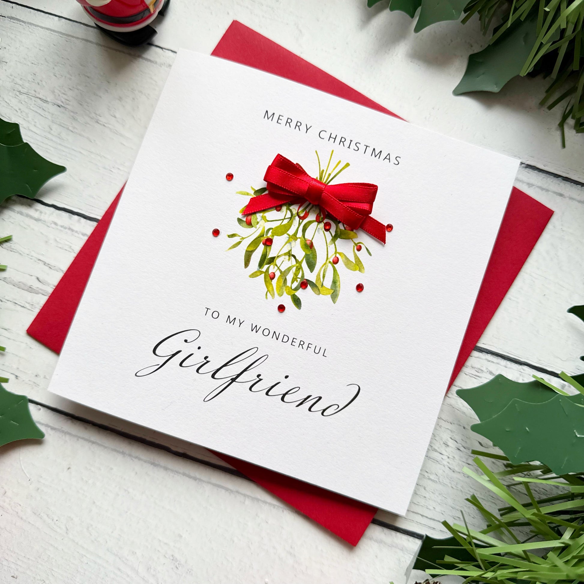 A square white Christmas card featuring a watercolour illustration of a mistletoe bunch with green leaves and red berries, tied with a real red satin ribbon bow and accented with small red gem embellishments scattered around the design. The text "Merry Christmas" is printed in small spaced capitals at the top of the card, with "to my Wonderful" in small capitals and "Girlfriend" in large elegant black cursive script below the illustration. The card sits on a matching red envelope on a white painted wooden s
