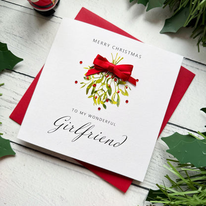 A square white Christmas card featuring a watercolour illustration of a mistletoe bunch with green leaves and red berries, tied with a real red satin ribbon bow and accented with small red gem embellishments scattered around the design. The text "Merry Christmas" is printed in small spaced capitals at the top of the card, with "to my Wonderful" in small capitals and "Girlfriend" in large elegant black cursive script below the illustration. The card sits on a matching red envelope on a white painted wooden s