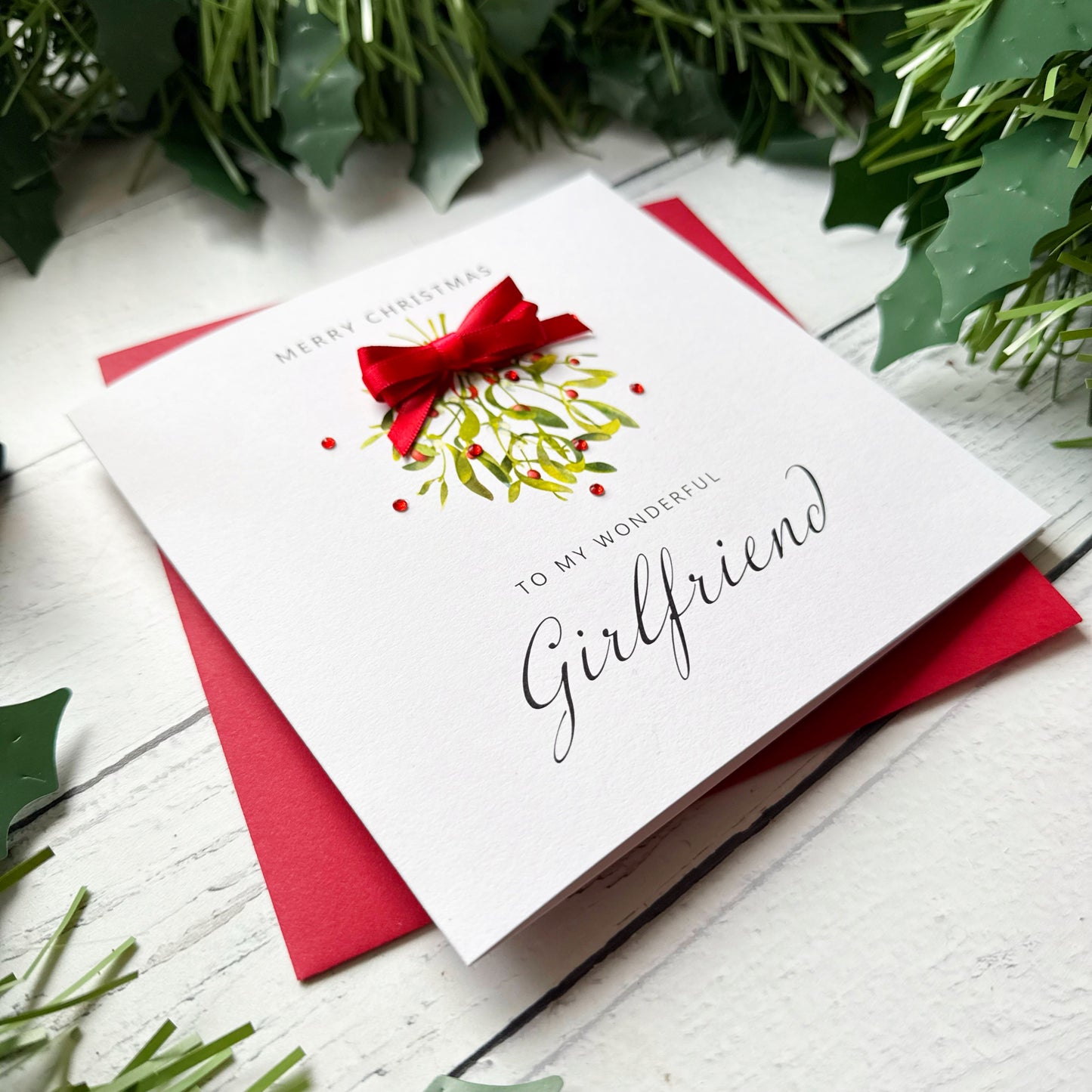 A white Christmas greeting card with a red bow and holly illustration, reading 'Merry Christmas to my wonderful Girlfriend,' placed on a red envelope over a white wood surface with green holly leaves around it.