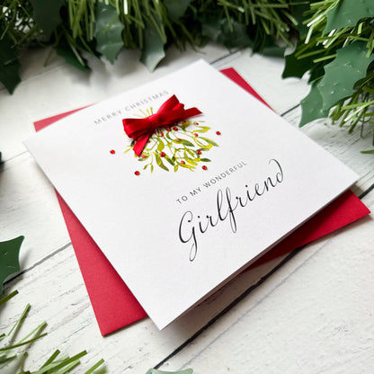 A white Christmas greeting card with a red bow and holly illustration, reading 'Merry Christmas to my wonderful Girlfriend,' placed on a red envelope over a white wood surface with green holly leaves around it.