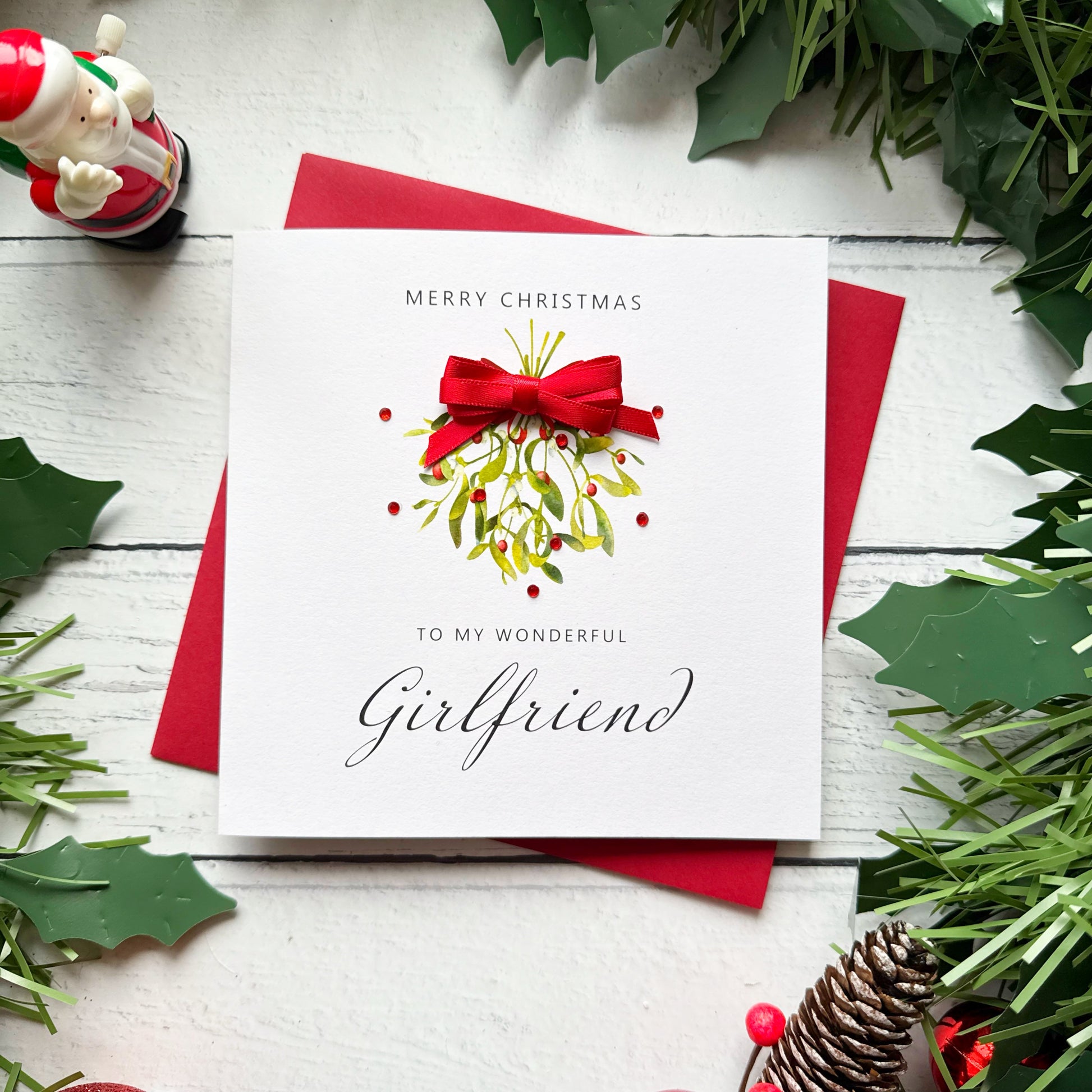 A square white Christmas greeting card with a red bow and mistletoe design, reading 'Merry Christmas to my wonderful Girlfriend,' placed on red envelopes on a white wood surface with holly and pine decorations around it.
