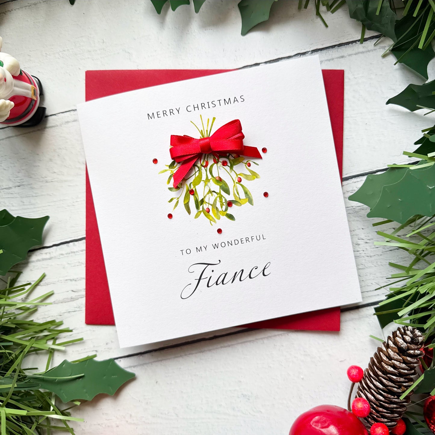 A square white Christmas greeting card with a red bow and mistletoe illustration, placed on top of a red envelope on a white wood surface with holiday greenery around it. The card reads, 'MERRY CHRISTMAS TO MY WONDERFUL Fiance.