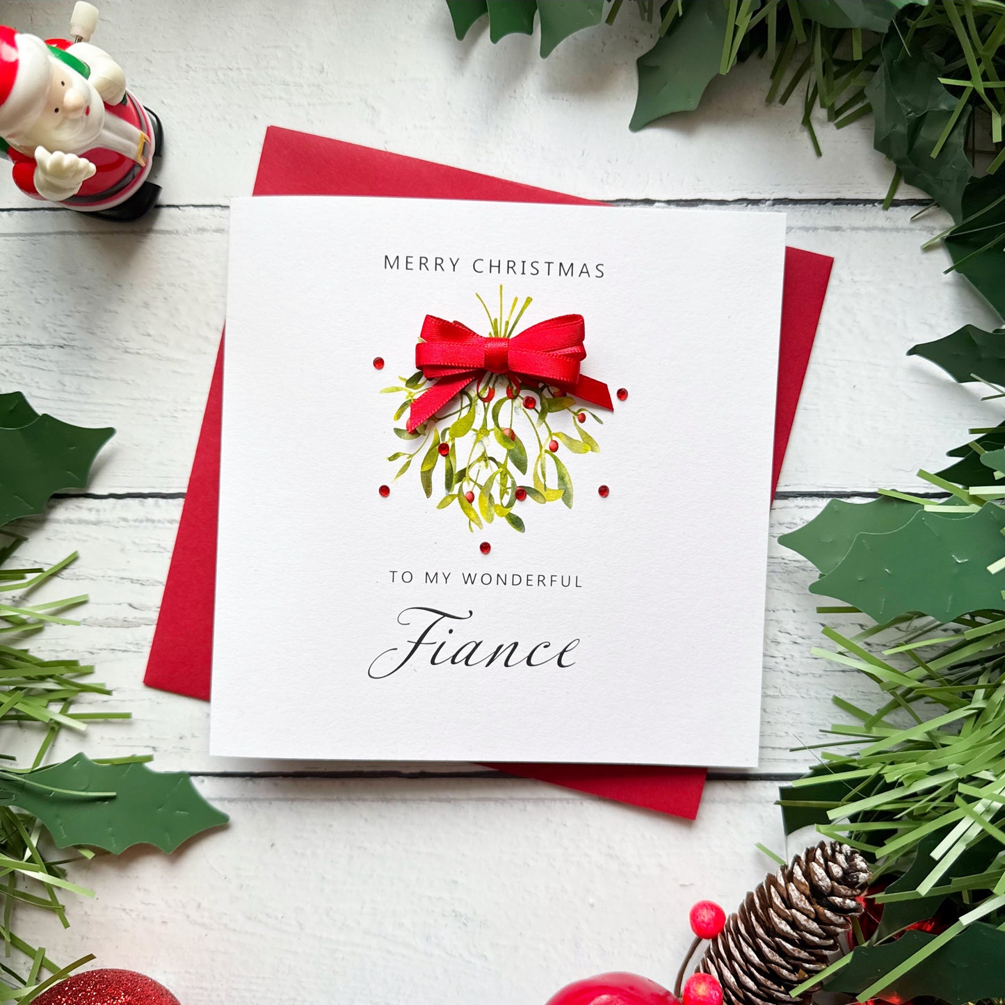 A square Christmas greeting card with a white front and red envelope, styled on a white wood surface with holly leaves, pinecones, and a small Santa figurine. The card reads “Merry Christmas to my wonderful Fiance” and features a red bow and green mistletoe illustration.