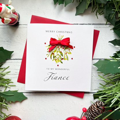 A square Christmas greeting card with a white front and red envelope, styled on a white wood surface with holly leaves, pinecones, and a small Santa figurine. The card reads “Merry Christmas to my wonderful Fiance” and features a red bow and green mistletoe illustration.