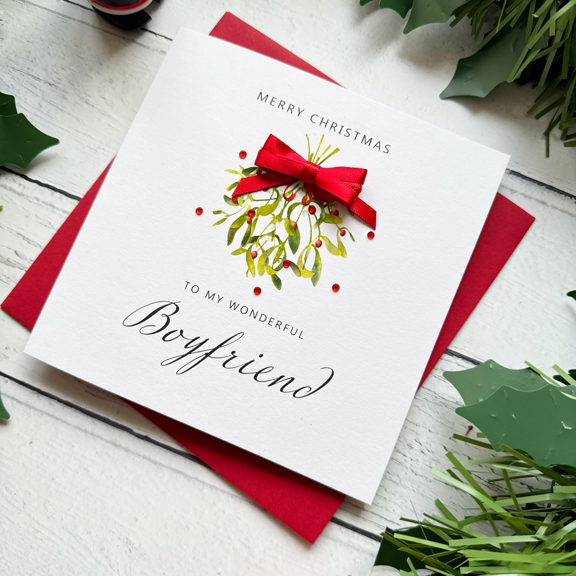 A square white Christmas greeting card with a red envelope, styled on a white wood surface with holly leaves and greenery. The card reads 'MERRY CHRISTMAS TO MY WONDERFUL Boyfriend' and features a red bow, mistletoe, and berries illustration.
