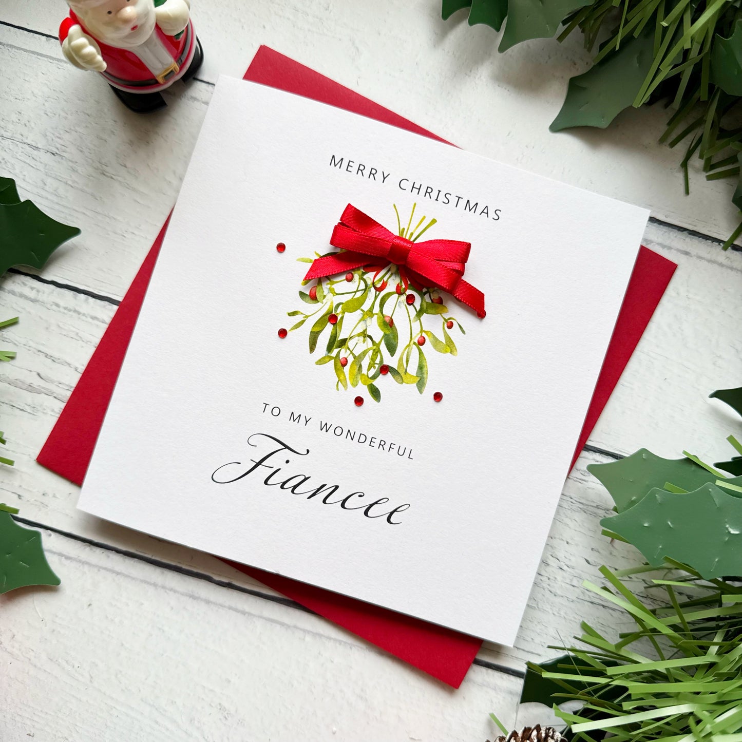 A white Christmas greeting card addressed 'To My Wonderful Fiancee' with a red bow and mistletoe illustration, placed on a red envelope on a white wood surface with holly leaves and a small Santa figurine around it.