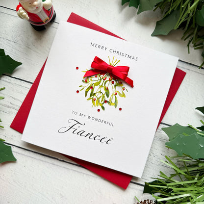 A white Christmas greeting card addressed 'To My Wonderful Fiancee' with a red bow and mistletoe illustration, placed on a red envelope on a white wood surface with holly leaves and a small Santa figurine around it.